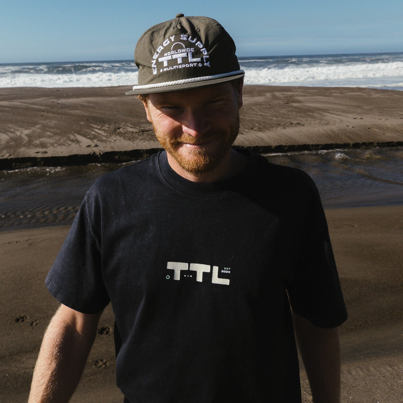 Man wearing a cap and black t-shirt with 'TTL' logo on a beach