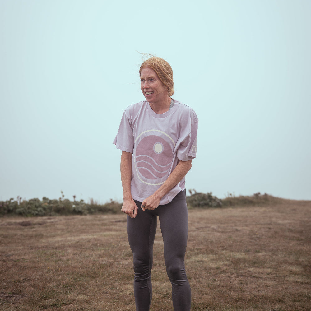 Person wearing a light purple t-shirt with a graphic design, standing in an open field.