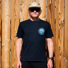 Man wearing a black t-shirt with a logo and sunglasses, standing in front of a wooden fence.