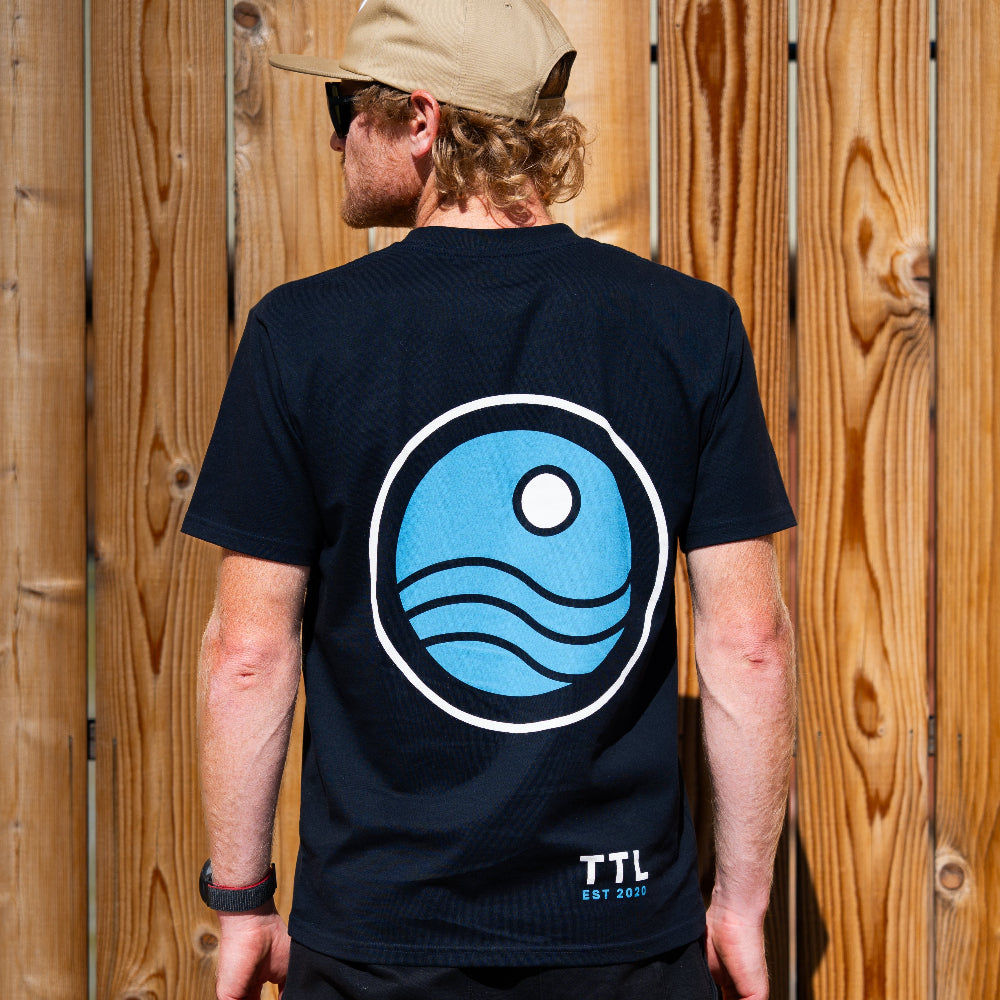 Person wearing a black t-shirt with a blue circular logo and 'TTL' branding, standing against a wooden fence.