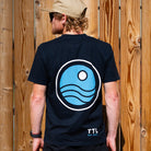 Person wearing a black t-shirt with a blue circular logo and 'TTL' branding, standing against a wooden fence.