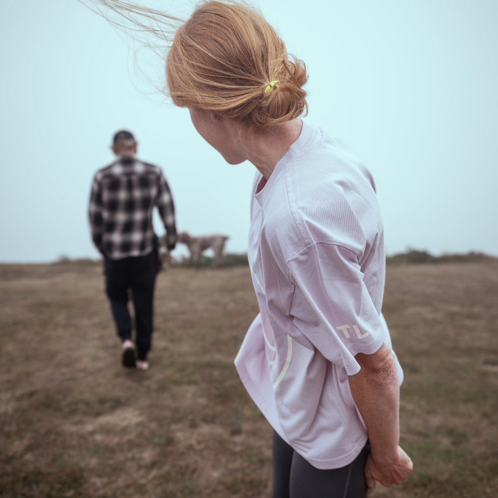 Person in a white shirt and dark pants standing on a grassy field with another person in the distance.