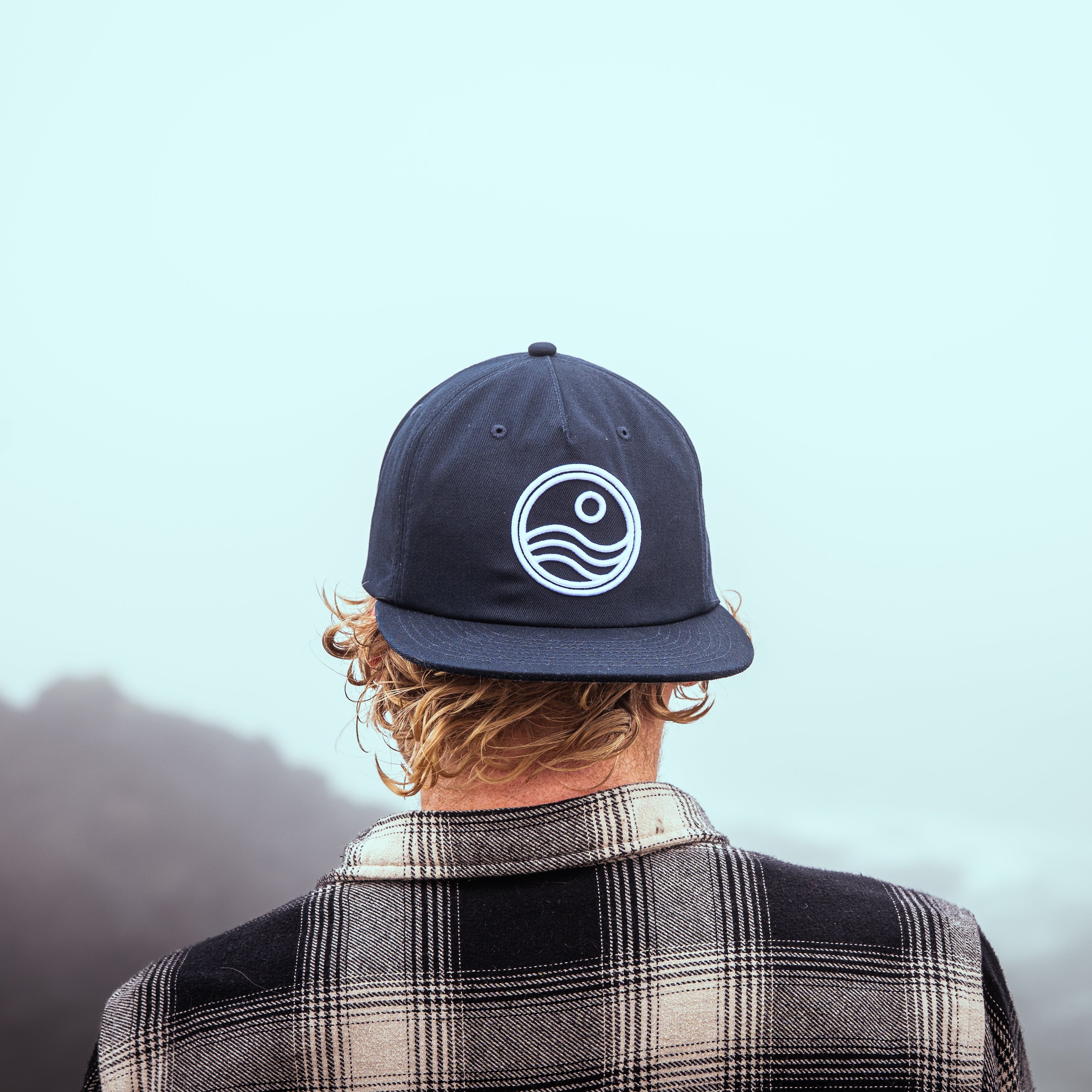 Person wearing a navy blue cap with a logo, facing away from the camera with a blurred natural background.