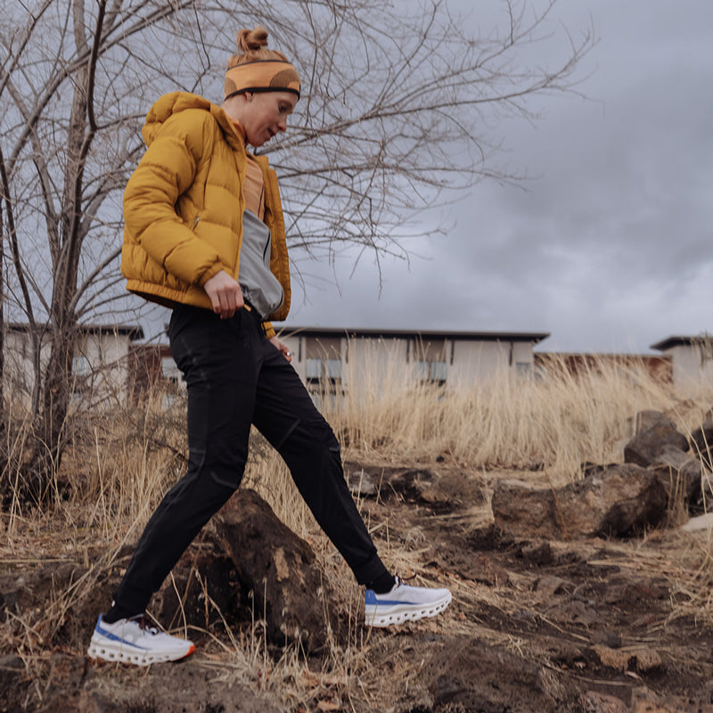 Person wearing On Cloudmonster shoes with a yellow jacket and black pants, walking through a rocky outdoor trail on a cloudy day.