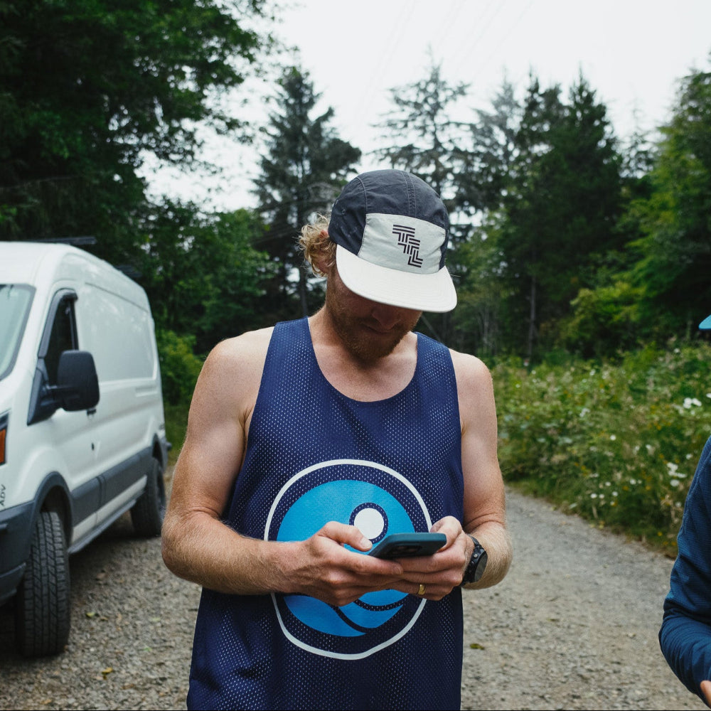 Two people standing outdoors, one using a phone, with a white van and trees in the background.