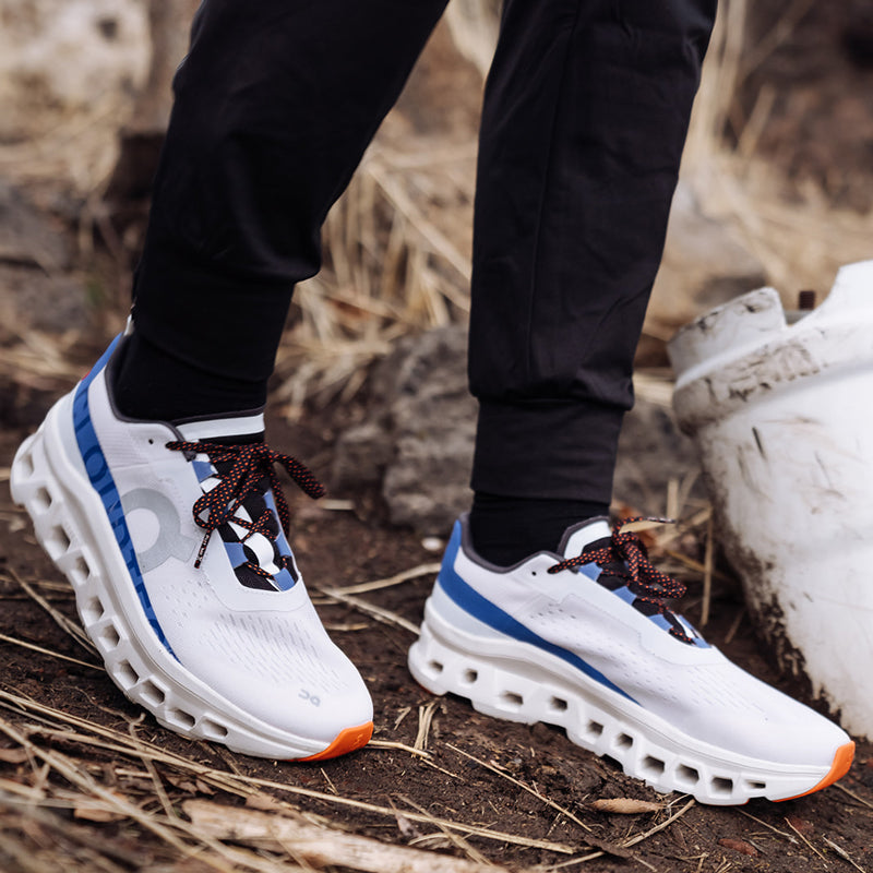 Close-up view of the On Cloudmonster shoes in white and blue, worn on a trail.