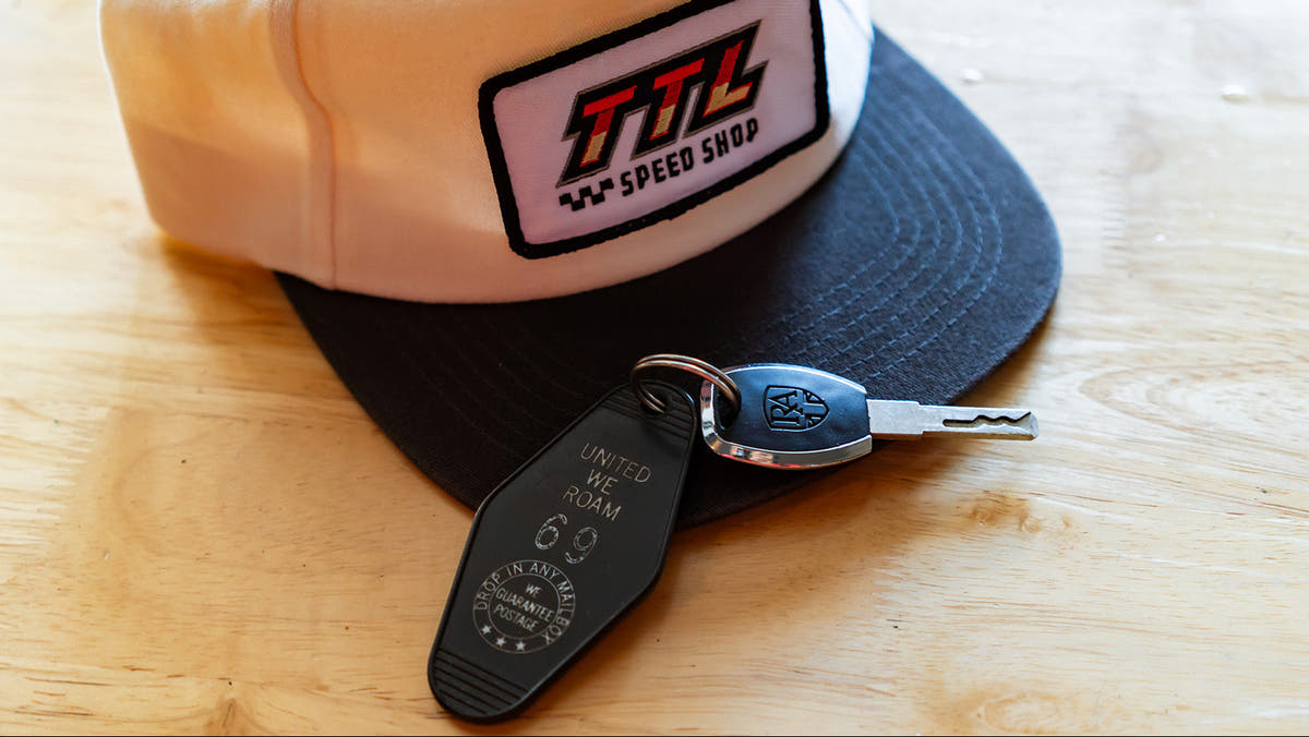A cream and black snapback cap with a 'TTL Speed Shop' patch on the front, resting on a light wood table next to a black keychain with a car key fob.